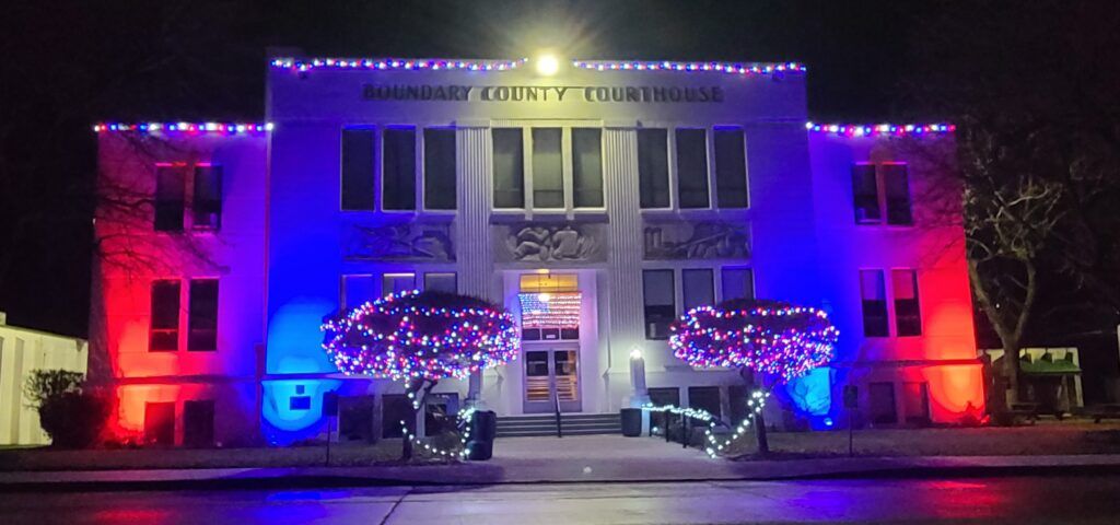 The Boundary County Courthouse in Boundary County ID shines at night with red, white, and blue lights, including lit trees and an American flag display in the entryway—a proud symbol of public service to the community. | Boundary County ID