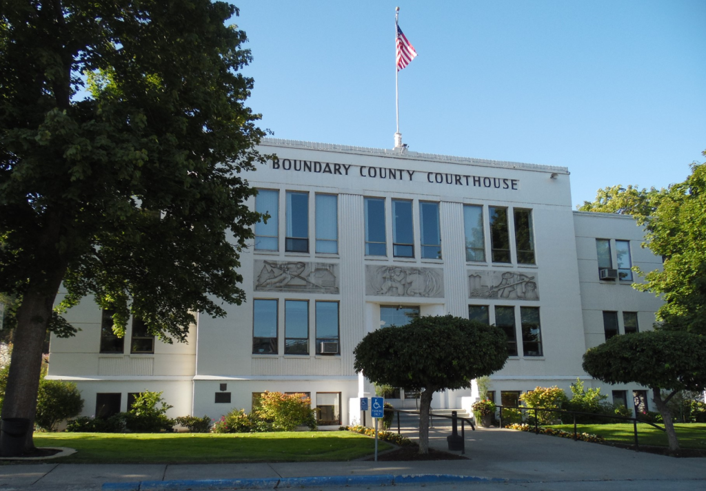 The Boundary County Courthouse in Boundary County ID is a white, two-story building with columns and sculpted panels, an American flag on the roof, trees in front, and a handicap parking sign visible in the foreground. | Boundary County ID