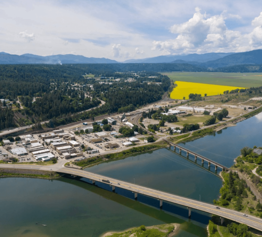 Aerial view of a river with two bridges in Boundary County ID, surrounded by a small town, forested hills, open fields, and mountains in the background under a partly cloudy sky. | Boundary County ID