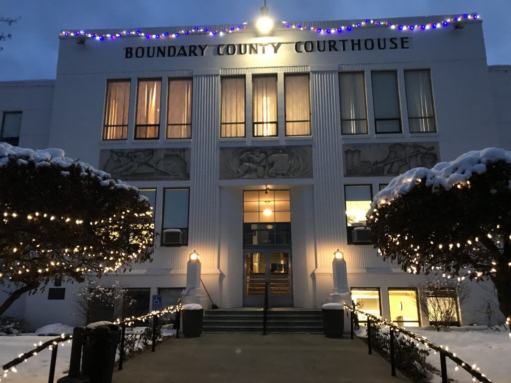 The Boundary County Courthouse in Boundary County ID is decorated with string lights and snow on the bushes and ground; illuminated windows, a lighted sign, and a sense of Public Service are visible in the evening. | Boundary County ID