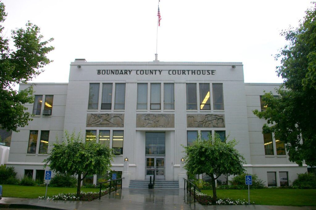 The two-story white Boundary County Courthouse in Boundary County ID features columns, large windows, decorative carvings, and an American flag on top, offering public service and public info with a landscaped, wheelchair-accessible entrance. | Boundary County ID