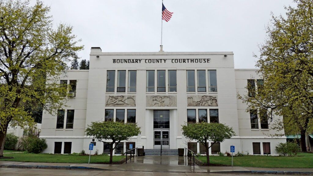 A white, three-story courthouse in Boundary County ID with "Boundary County Courthouse" on the front, an American flag atop the roof, trees on each side, accessible parking in front—serving as a hub for public info and public service. | Boundary County ID