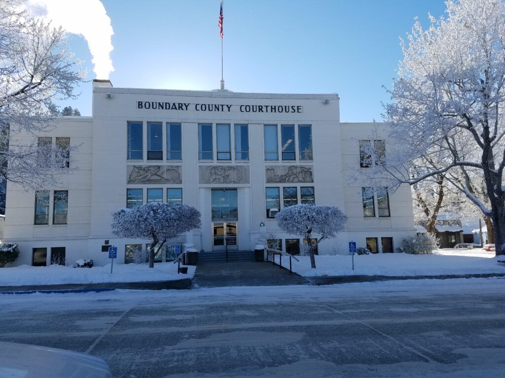 A white, three-story building labeled "Boundary County Courthouse" stands amid snow-covered ground and trees in Boundary County ID, proudly serving the public. An American flag waves on the roof beneath a clear, bright blue sky. | Boundary County ID