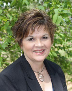 A woman with short, brown hair is smiling at the camera. She is wearing a black blazer, light earrings, and a circular pendant necklace. Green foliage is visible in the background, reflecting her dedication to Public Service in Boundary County ID. | Boundary County ID