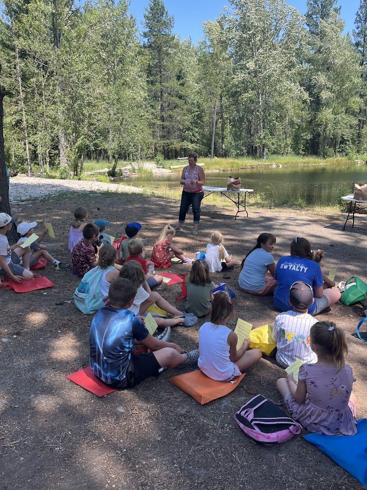 A group of children in Boundary County, ID sit on mats outdoors, listening attentively to a public service presentation near a pond and trees. They hold papers while tables with items are set up nearby. It is a sunny day. | Boundary County ID