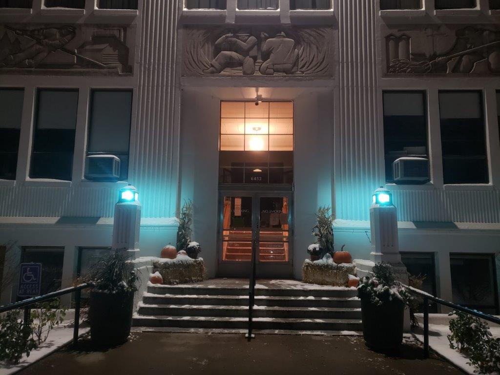 A building entrance at night in Boundary County ID features glowing blue-green lamps, steps dusted with snow, and seasonal pumpkins and hay bales on either side—an inviting scene for any public service or public info event. | Boundary County ID