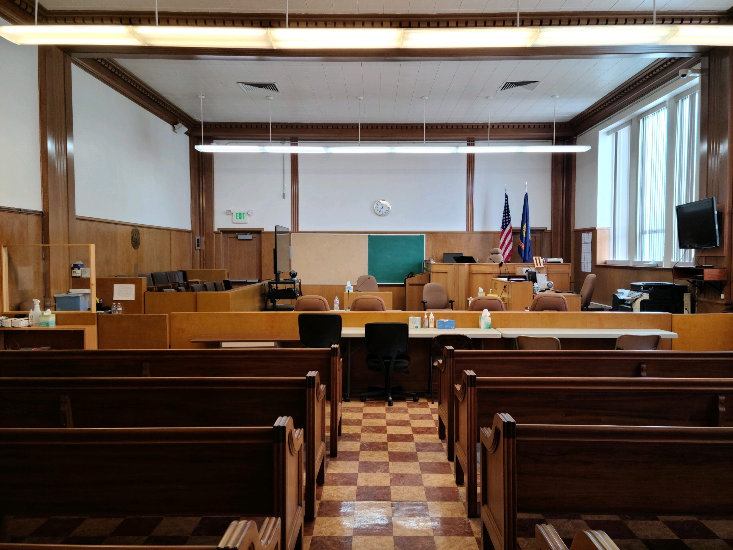 Empty Boundary County ID courtroom with wooden benches in the foreground, a checkered floor, judge’s bench, and desks. U.S. and state flags emphasize its role as a center for public service and access to public info. | Boundary County ID