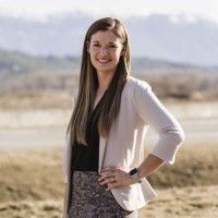 A woman with long straight hair stands outdoors in Boundary County ID, smiling at the camera. She wears a light-colored blazer, dark top, and patterned skirt, ready to share public info against a beautiful mountain backdrop. | Boundary County ID