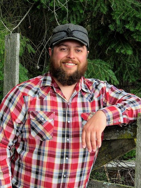 A bearded man wearing a gray cap and a red plaid shirt stands outdoors in Boundary County ID, leaning on a wooden fence with trees and greenery in the background. He is smiling and looking at the camera. | Boundary County ID