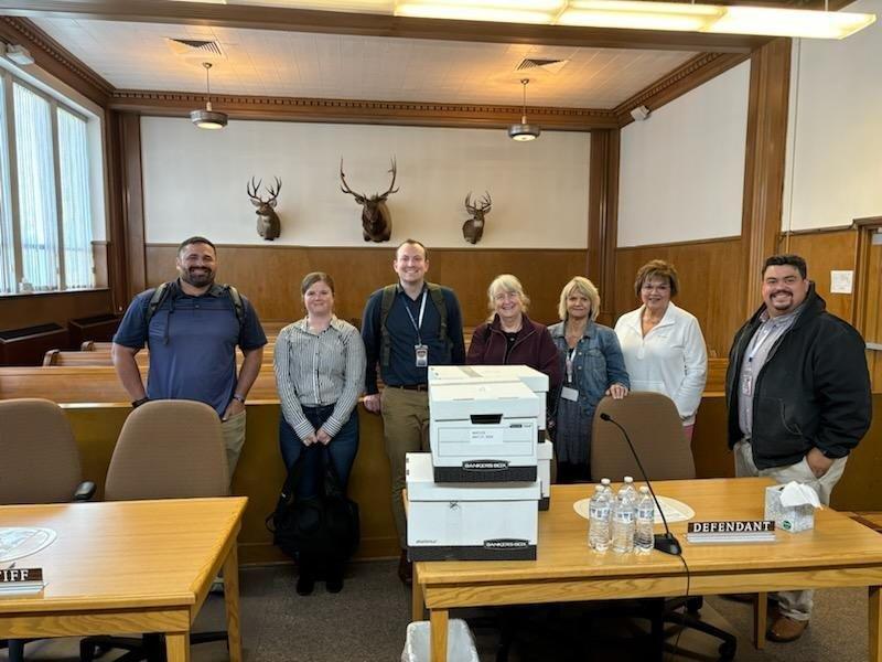 Seven people stand in a Boundary County ID courtroom behind a table with stacked boxes, bottled water, and nameplates labeled "Plaintiff" and "Defendant." Three deer heads are mounted on the wood-paneled wall behind them. | Boundary County ID