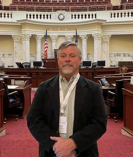 A man with gray hair and a goatee stands indoors wearing a black blazer and a conference badge, representing Public Service. Behind him is a government chamber with desks, computers, American flags, and "In God We Trust" on the wall. | Boundary County ID