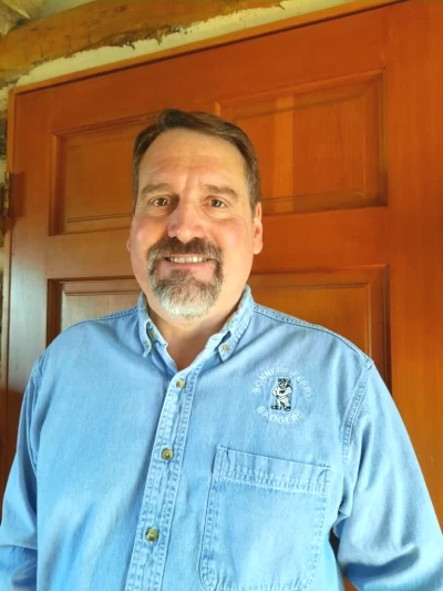 A middle-aged man with short brown hair, a mustache, and a goatee stands indoors in front of a wooden door, wearing a light blue button-up shirt with an embroidered Boundary County ID Public Service logo on the left side. | Boundary County ID