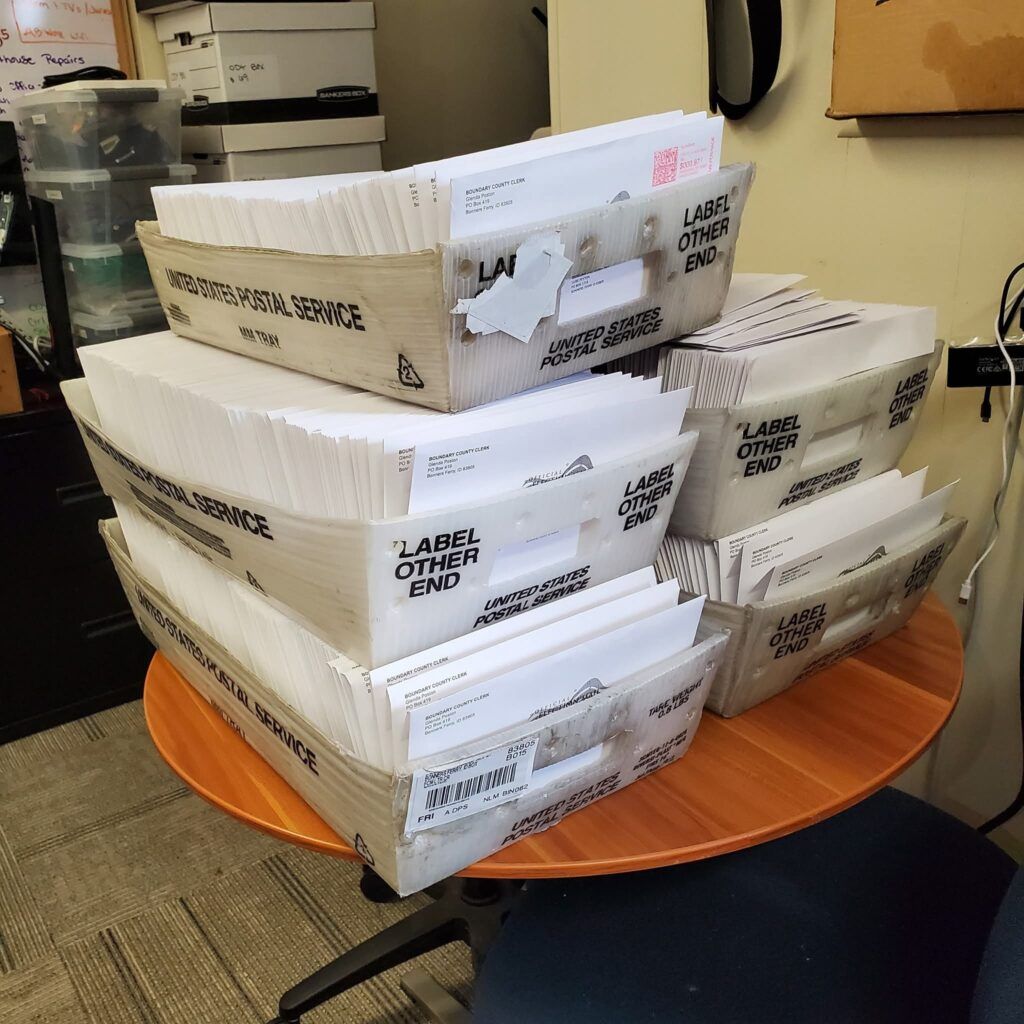 A round wooden table in a Boundary County ID office holds six large trays of sorted white envelopes labeled "United States Postal Service," with shelves and equipment visible in the background, ready for public service and information delivery. | Boundary County ID