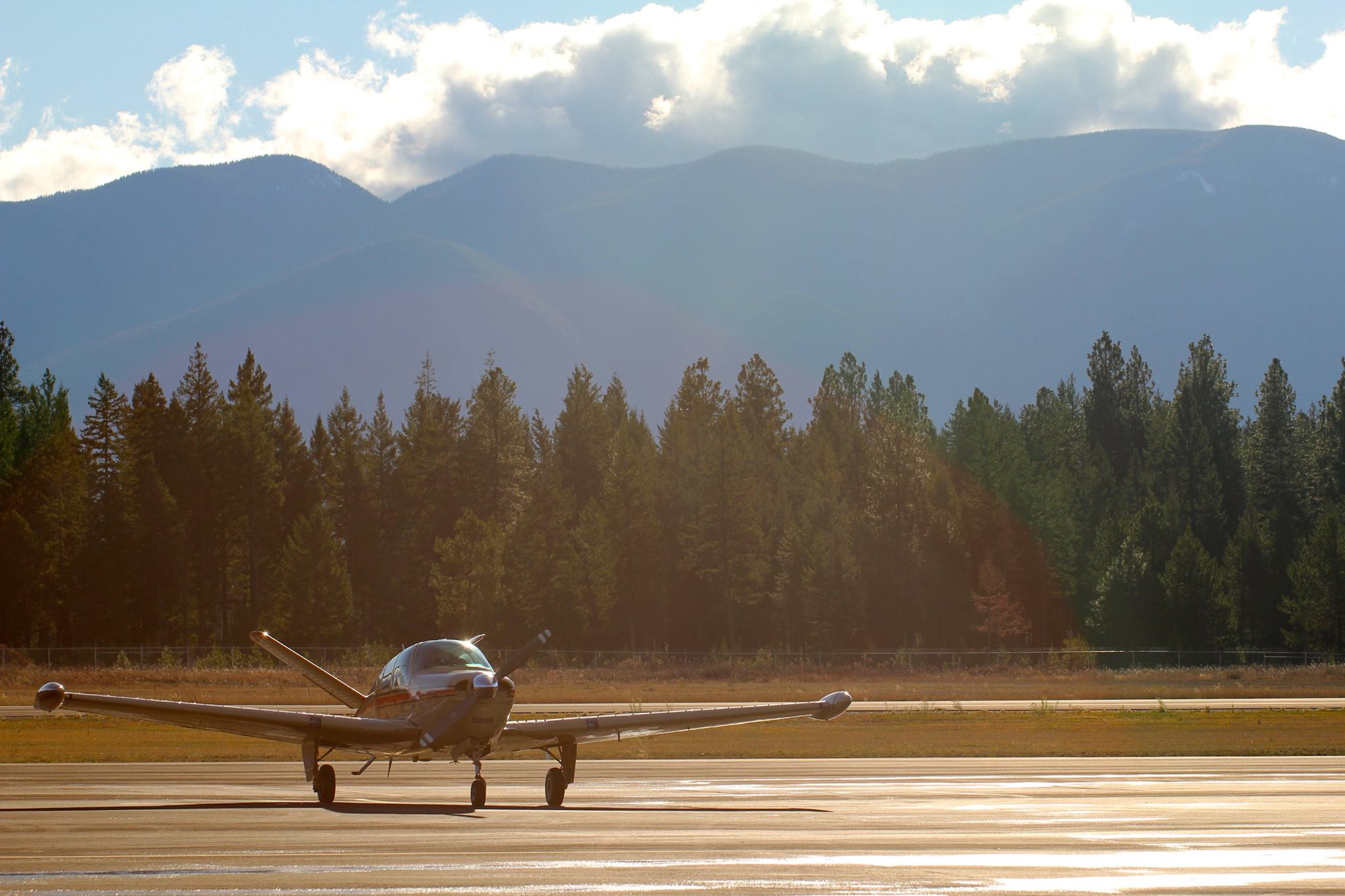 A small single-engine airplane is parked on a sunlit runway at Boundary County ID, with a line of trees and mountains in the background under a partly cloudy sky. | Boundary County ID